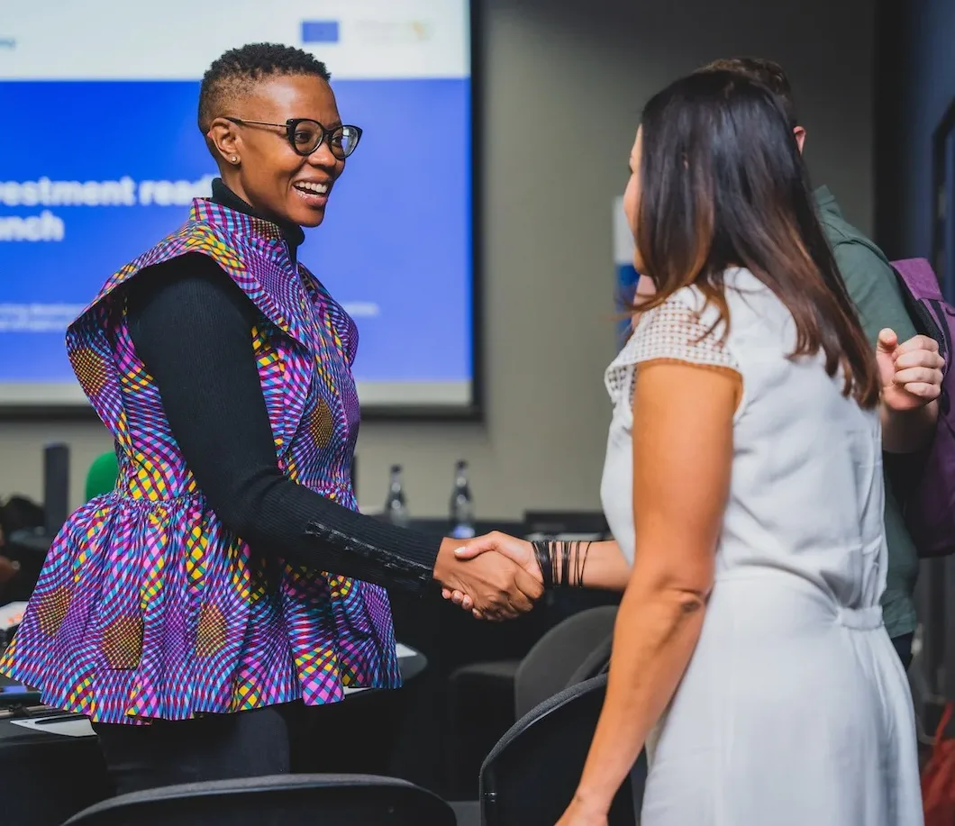 A medium shot of two smiling women of different races shaking hands as they meet for the first time at a corporate event covered by The Chalk Factory productions corporate event photography rates packages.